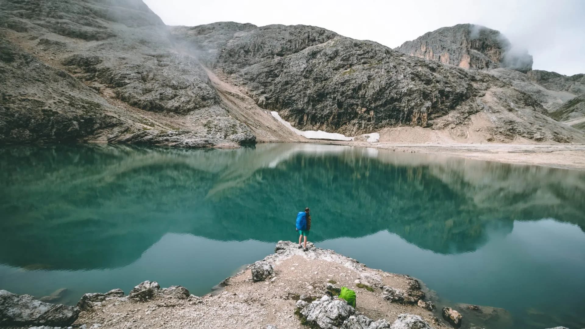 Hiker with backpack standing by turquoise alpine lake reflecting rocky mountains at Lago Antermoia.