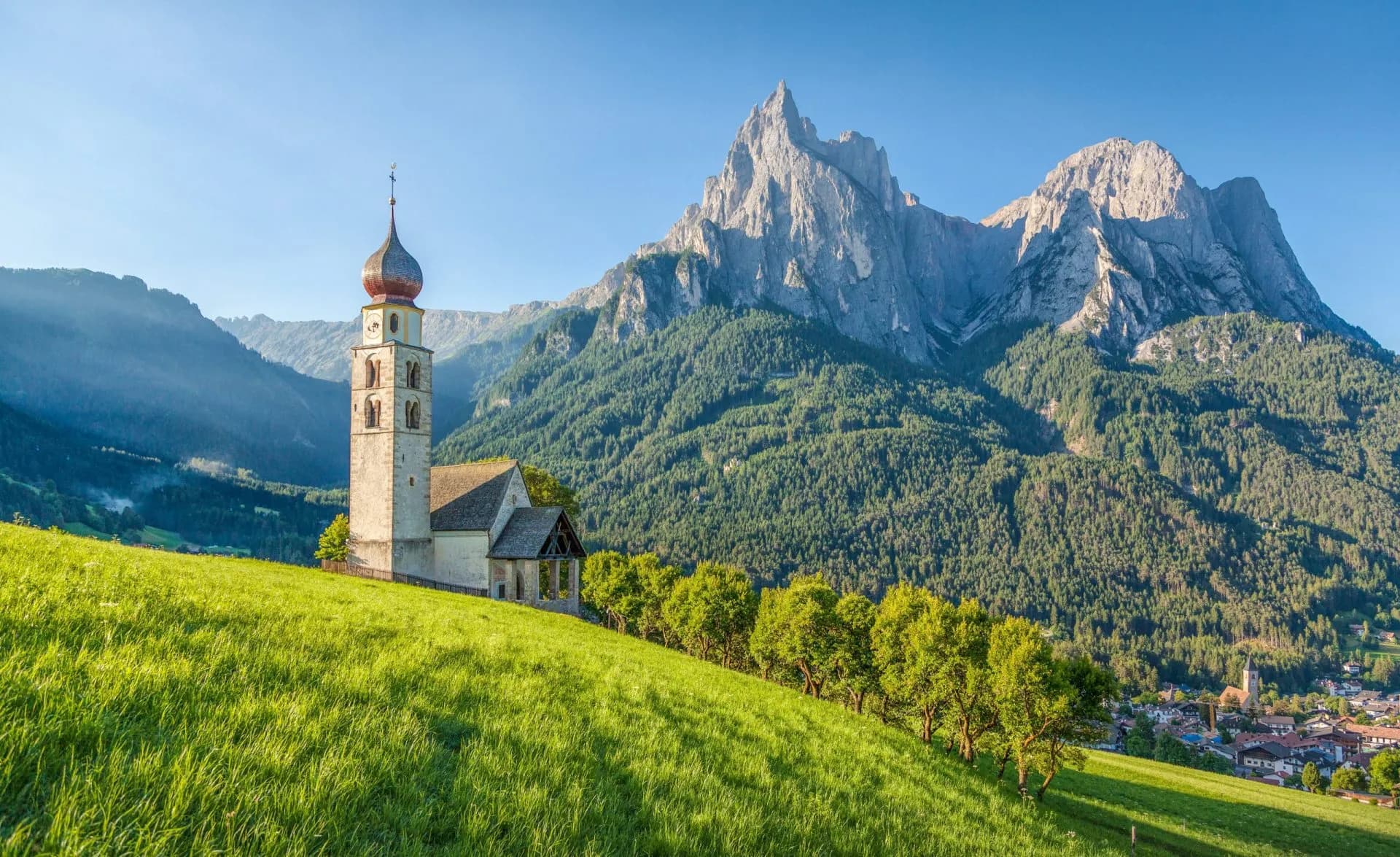 Church tower on grassy hill overlooking alpine village with dramatic mountain backdrop in Sëlva di Val Gardena.