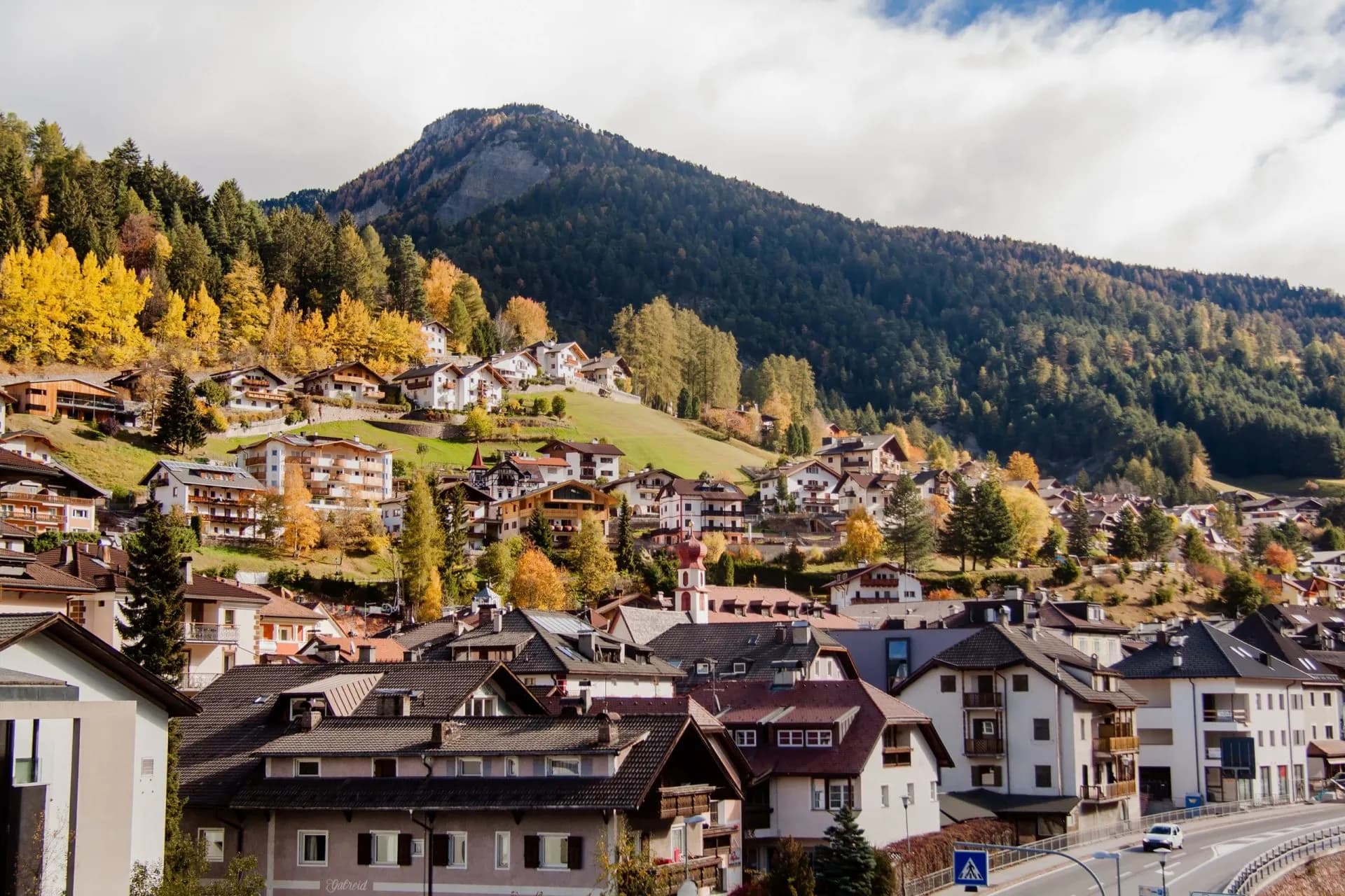 Alpine village houses climbing a steep hillside with autumn trees below a dark, forested mountain.