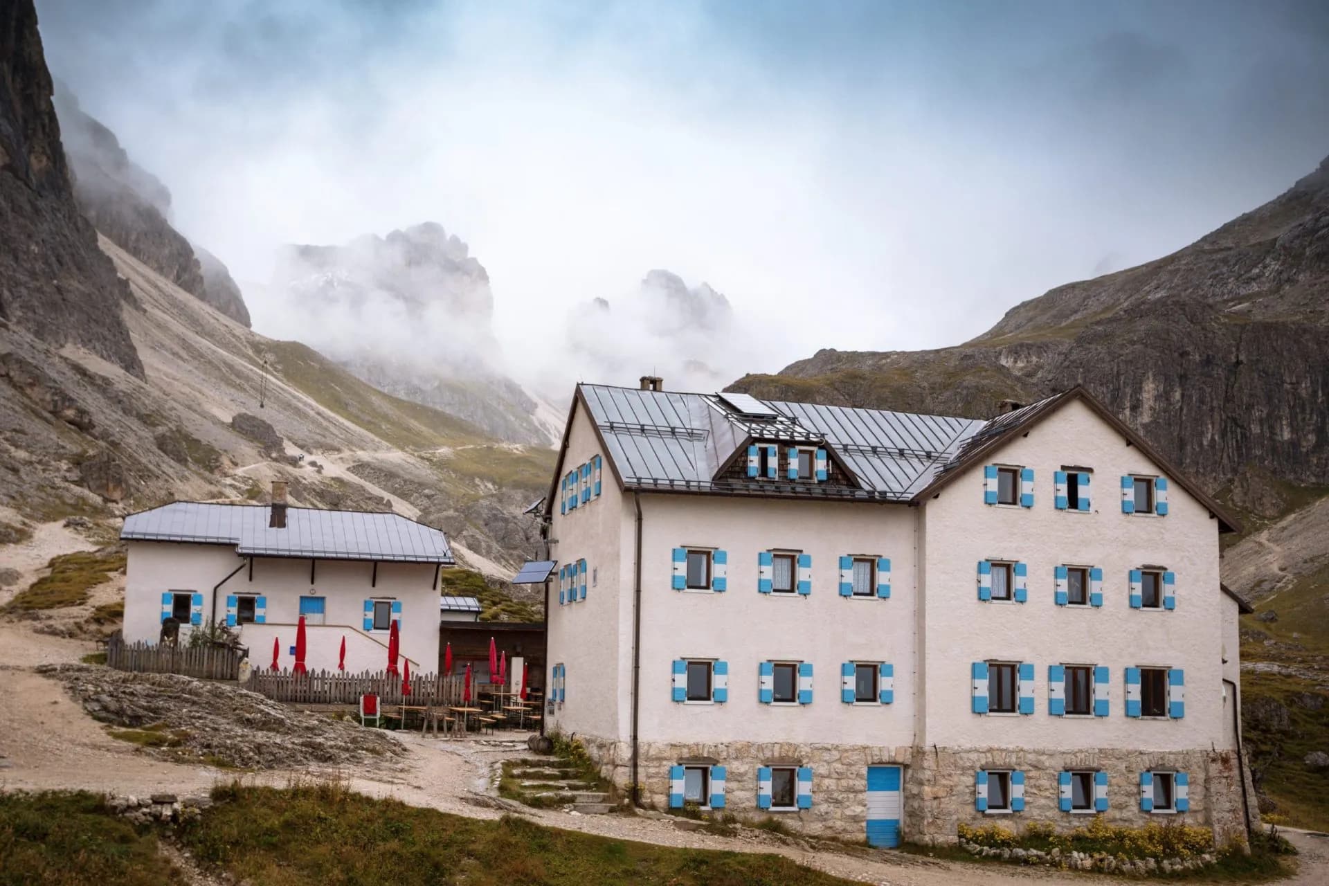 Rifugio Vajolet mountain hut with white walls and blue shutters nestled in foggy, rocky alpine terrain.