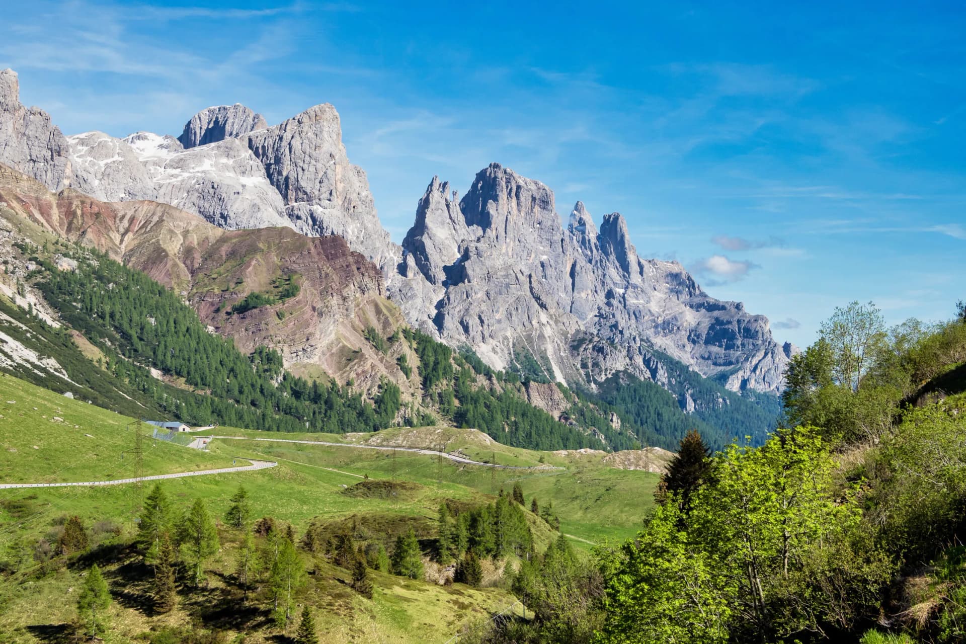 Pale di San Martino Dolomites from Passo Rolle with green slopes and winding road under blue sky.