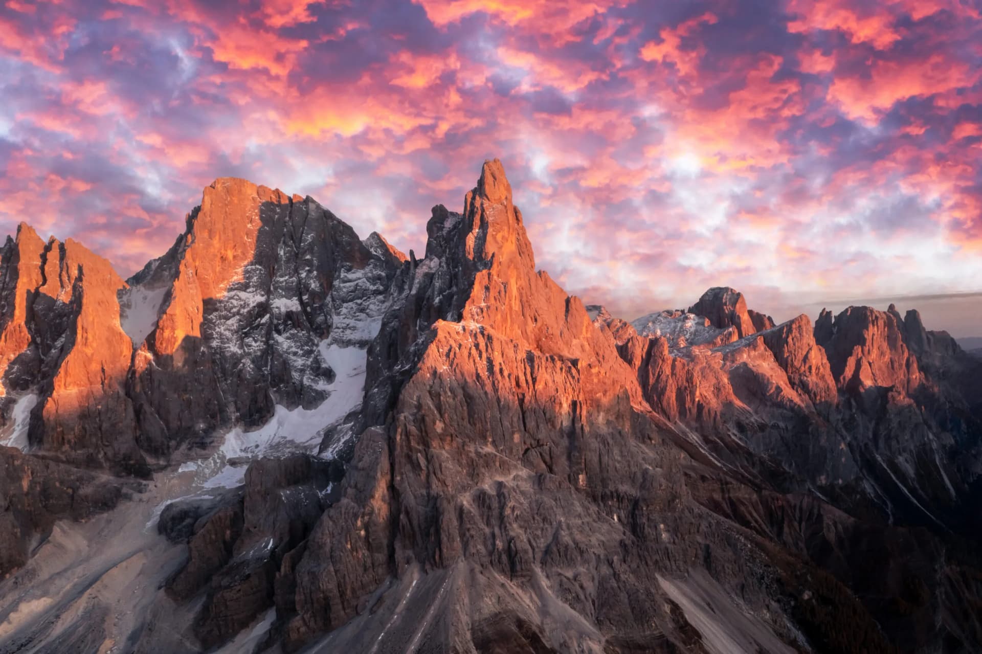 Rugged mountain peaks catching sunset light with snow patches under dramatic pink and purple clouds, Pale di San Martino.