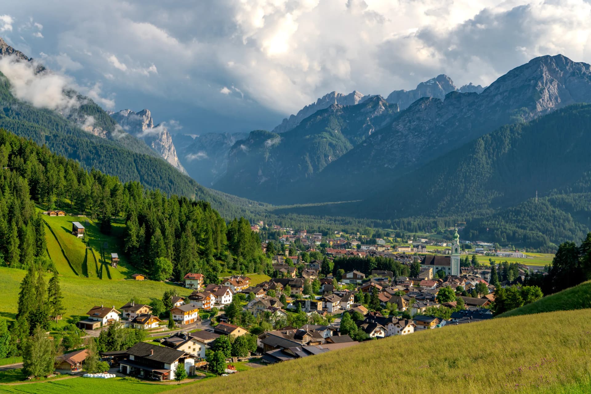 Alpine village nestled in valley with steep green slopes and dramatic mountains under cloudy sky, Dobbiaco.