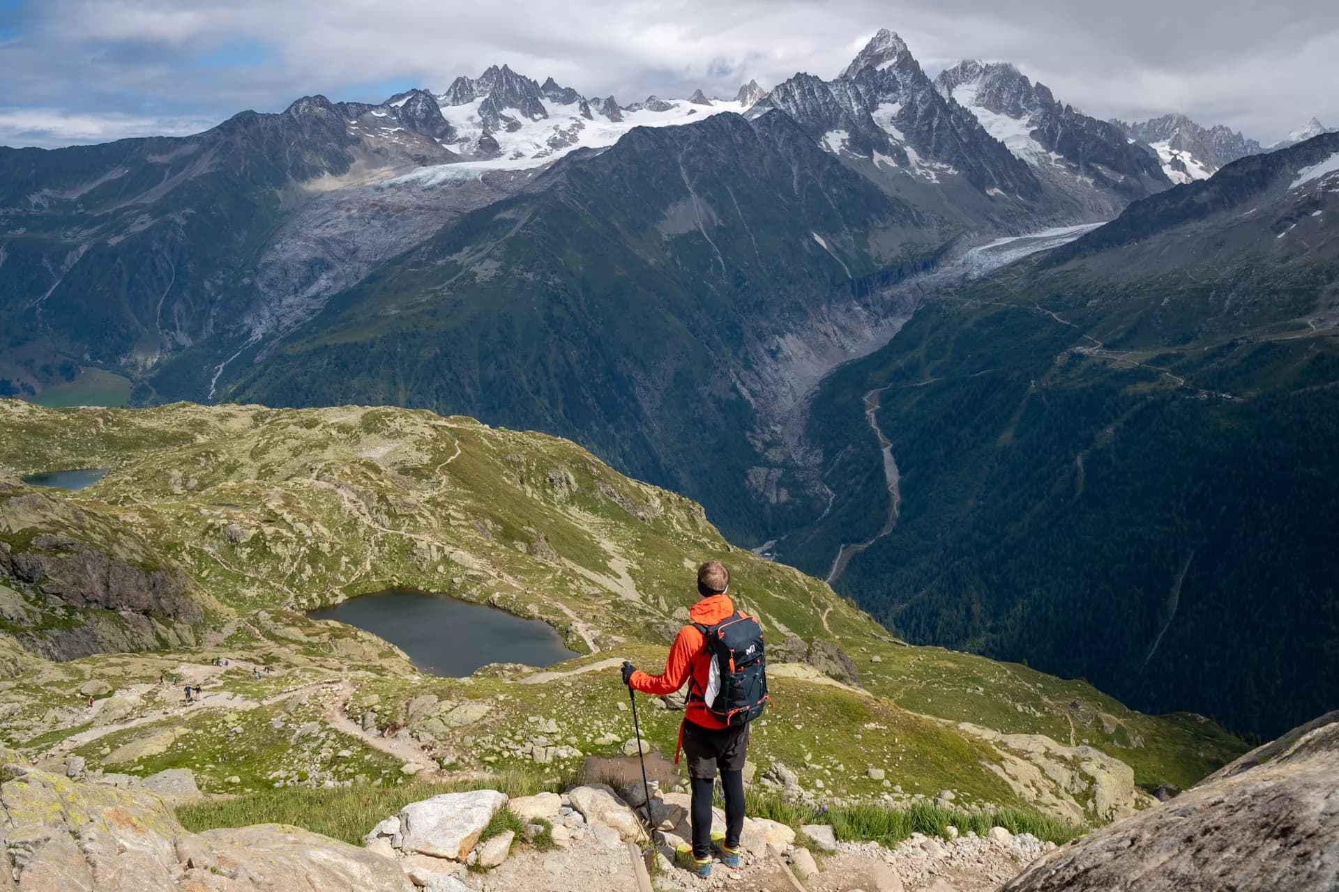 Hiker with backpack and poles overlooking alpine lakes and snow-capped mountains near Lacs de Cheserys.
