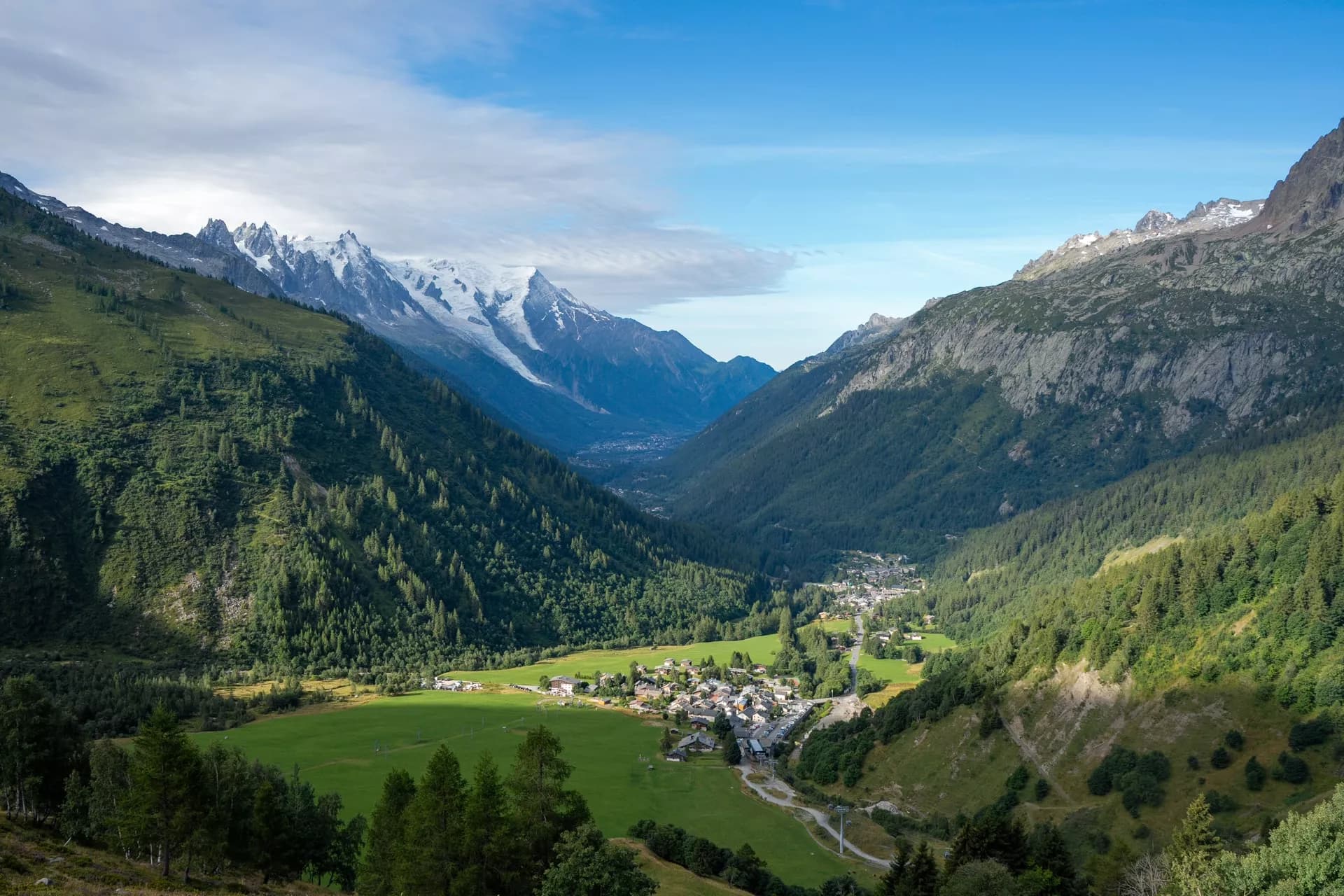Alpine valley with village, green slopes, and snow-capped mountains under blue sky, leaving Chamonix Valley.