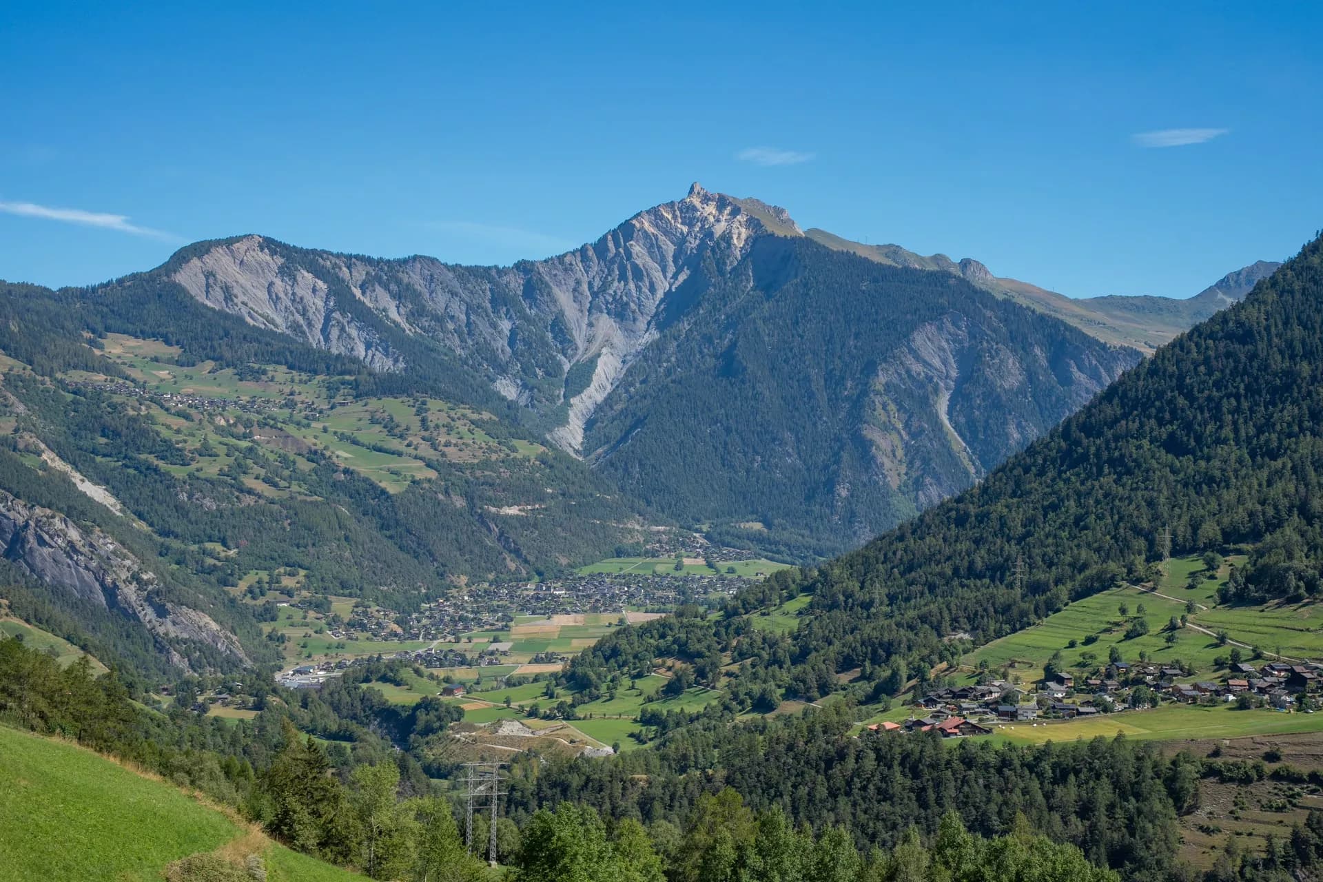Alpine valley with Val-de-Bagnes village nestled between steep, forested mountains under a clear blue sky.