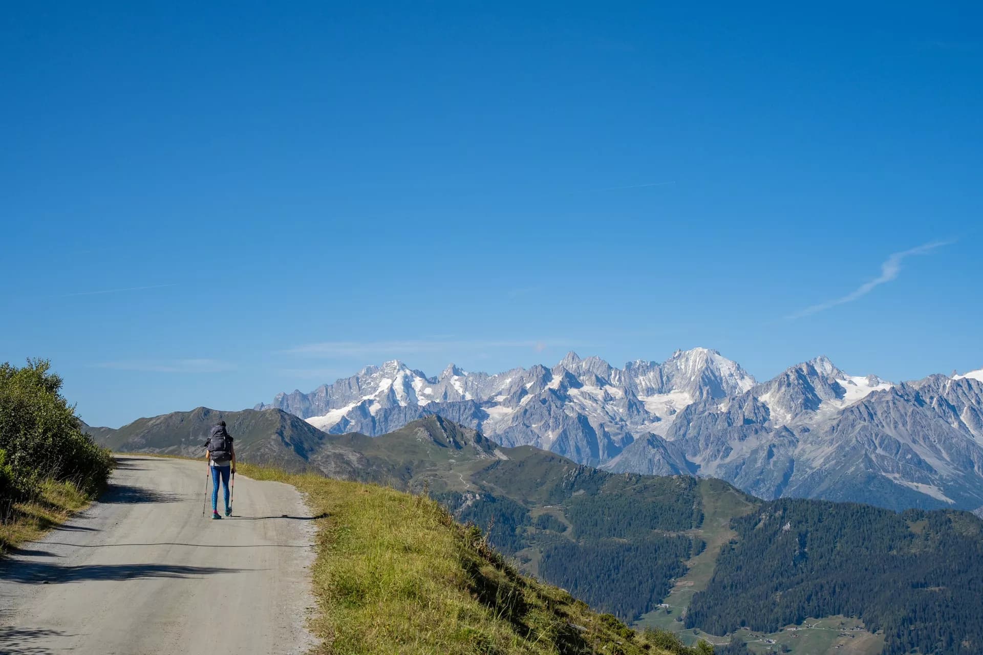 Hiker with backpack walking on dirt road toward snow-capped Mont Blanc Massif mountains.