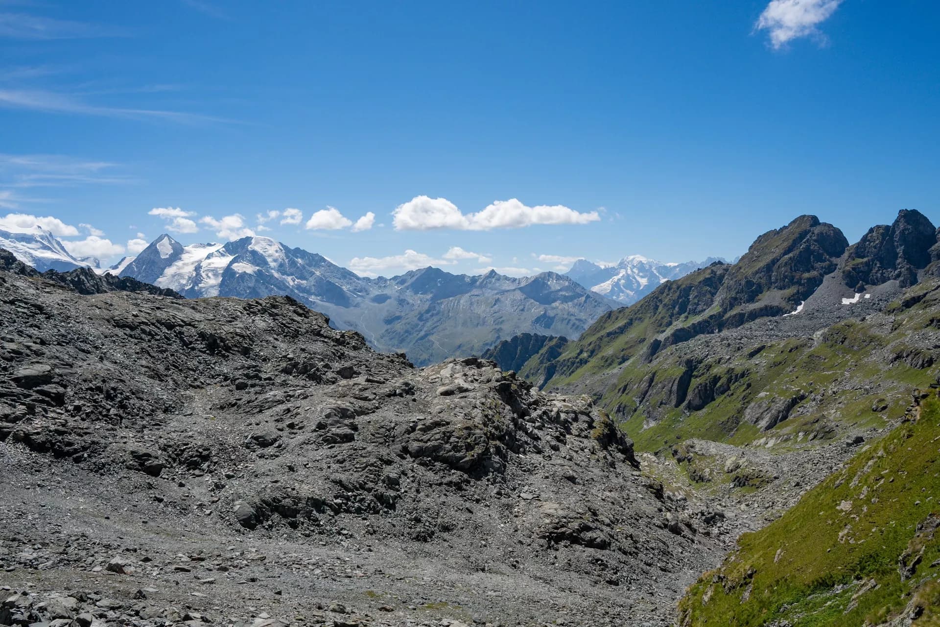 Rocky mountain terrain with green slopes and distant snow-capped peaks under a blue sky.