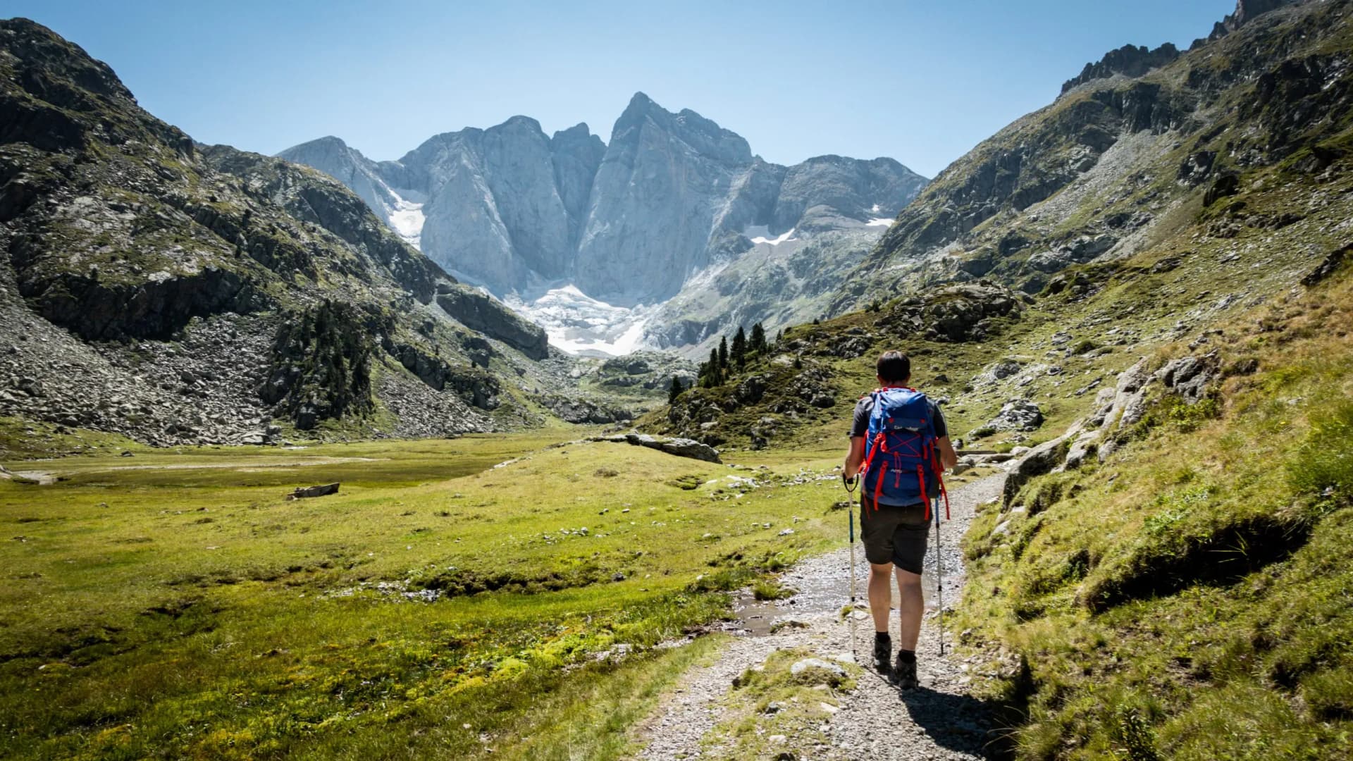 Hiker with backpack walking on trail toward massive rocky mountains and glacier remnants.