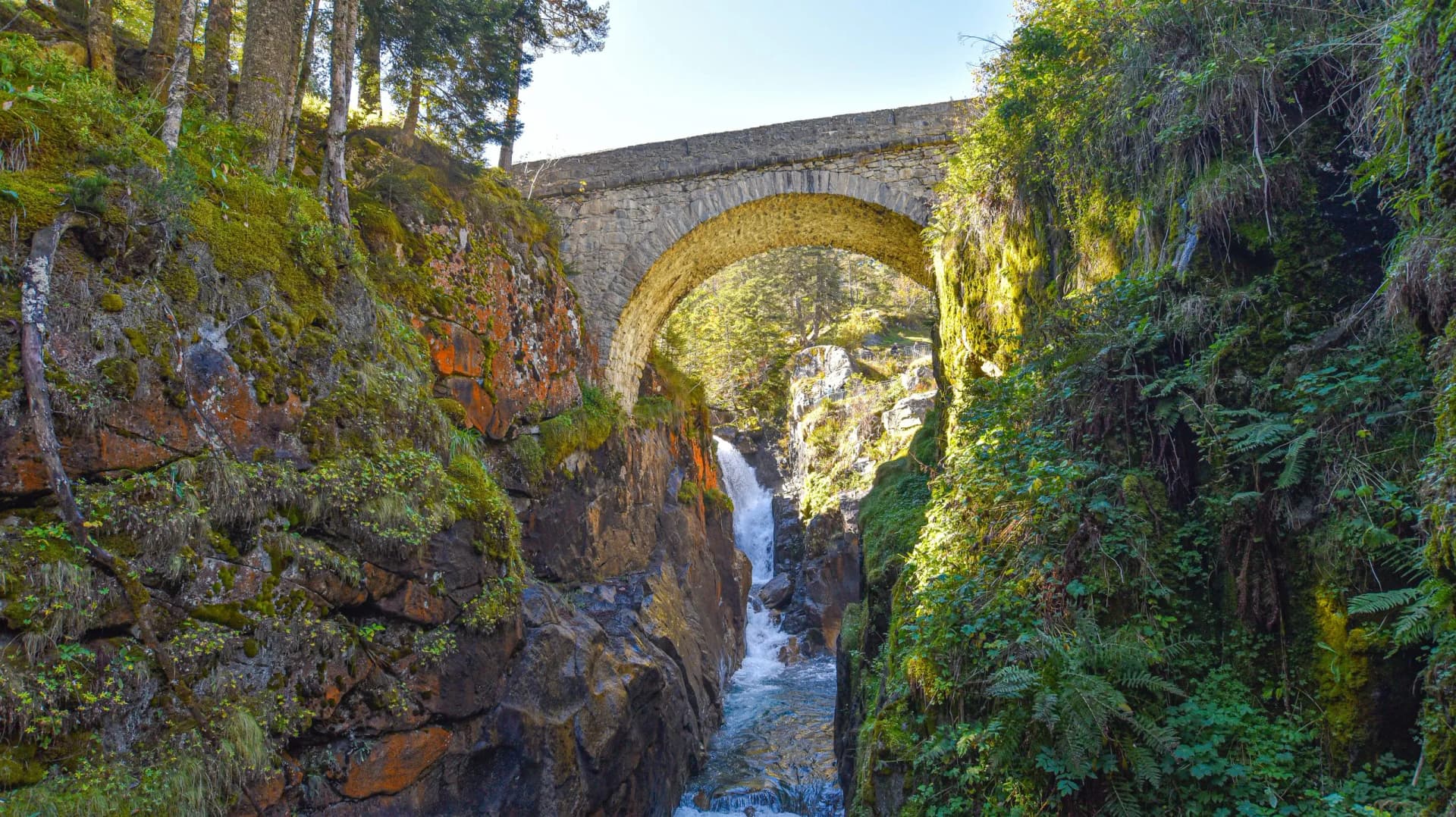 Stone arch bridge over a narrow gorge with a waterfall and lush green mossy banks