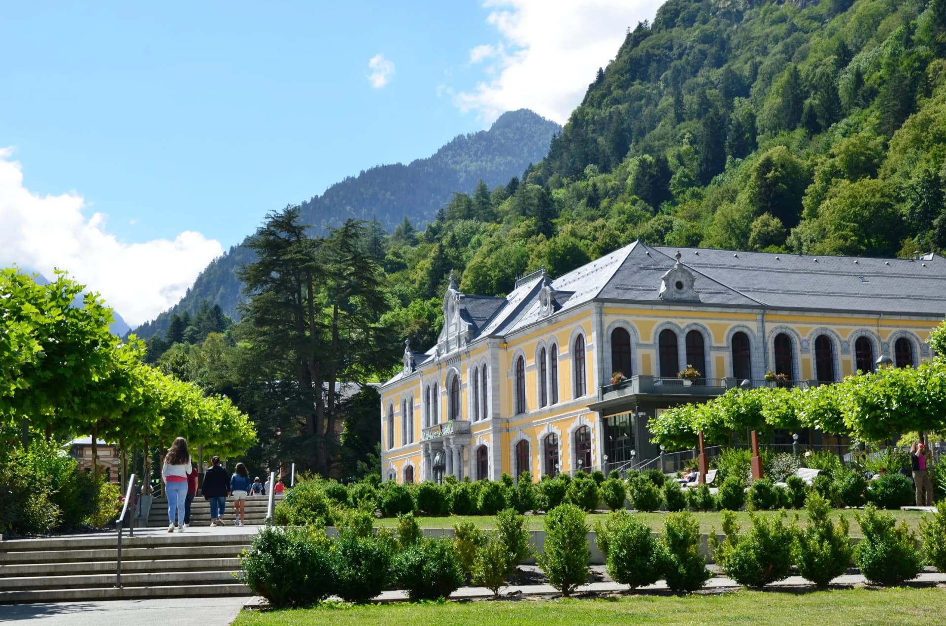 Yellow historic building in Cauterets park with forested mountains under blue sky.