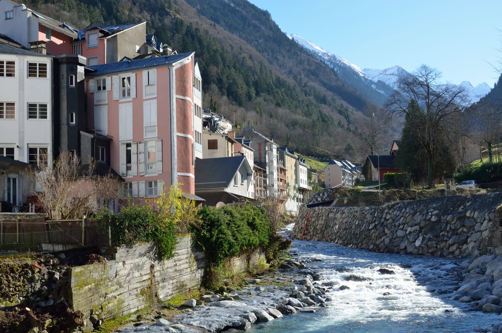Winter view of Cauterets spa town with buildings along a rocky, fast-flowing river and snow-capped mountains.