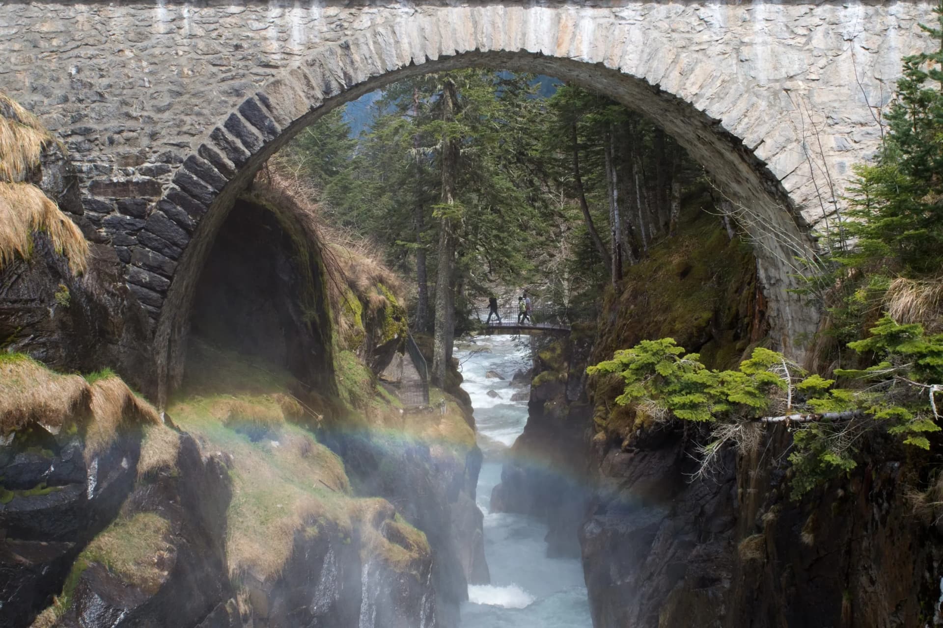 Stone arch bridge over a gorge with a river, hikers, and a rainbow.