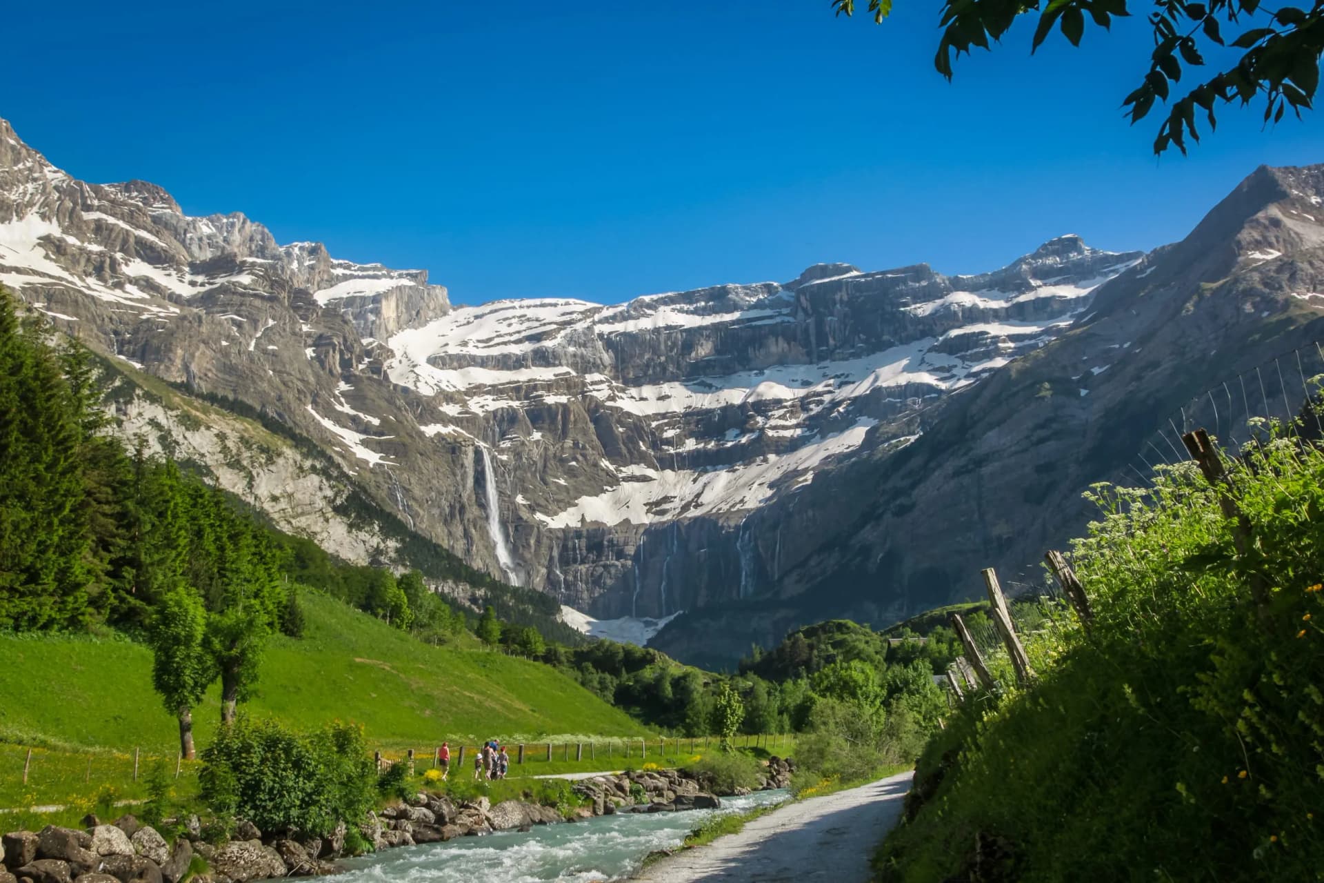 Cirque de Gavarnie with snow-capped mountains, a tall waterfall, and hikers by a rushing river.