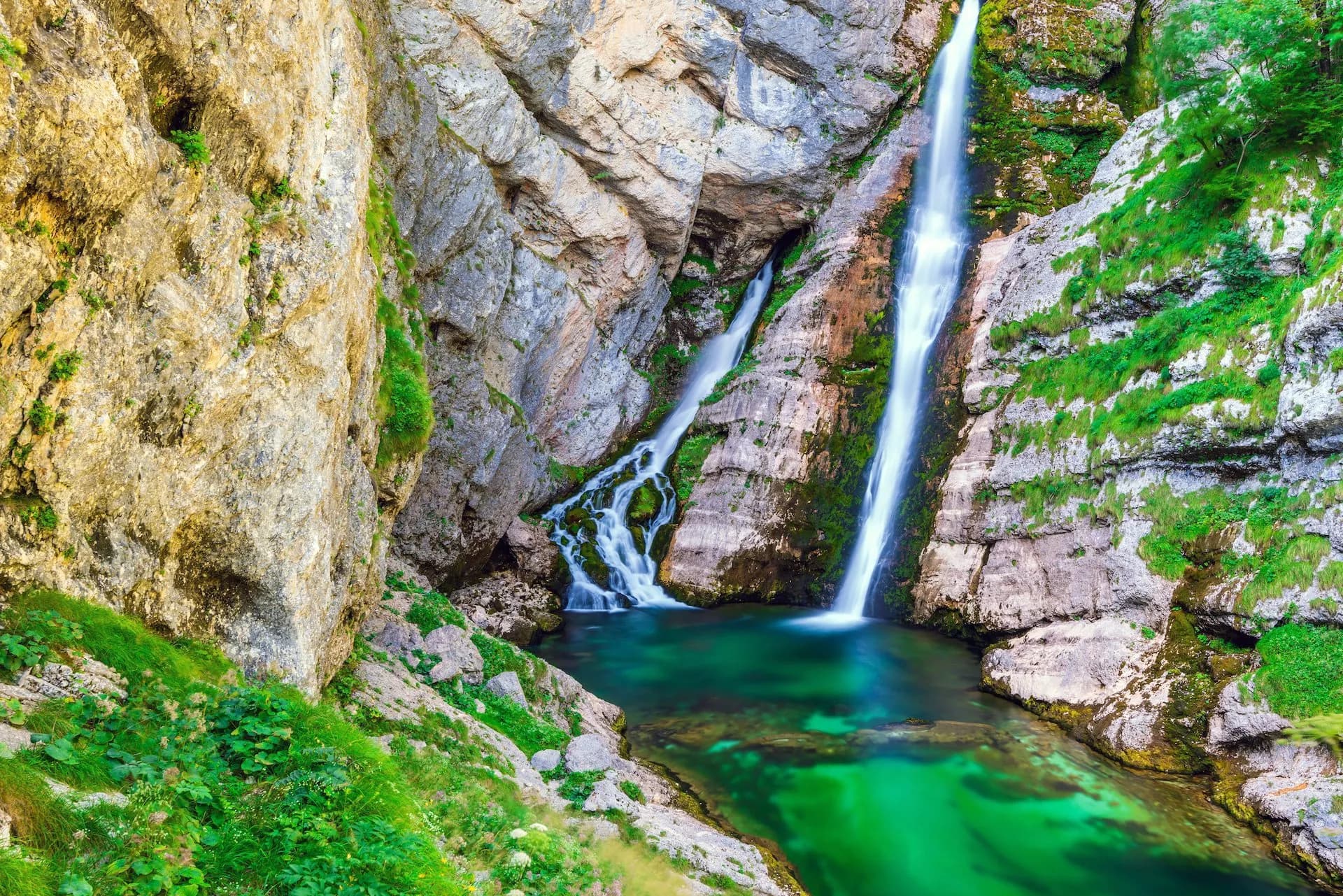Savica Waterfall cascading down rocky cliffs into a bright green pool in a mountain gorge.