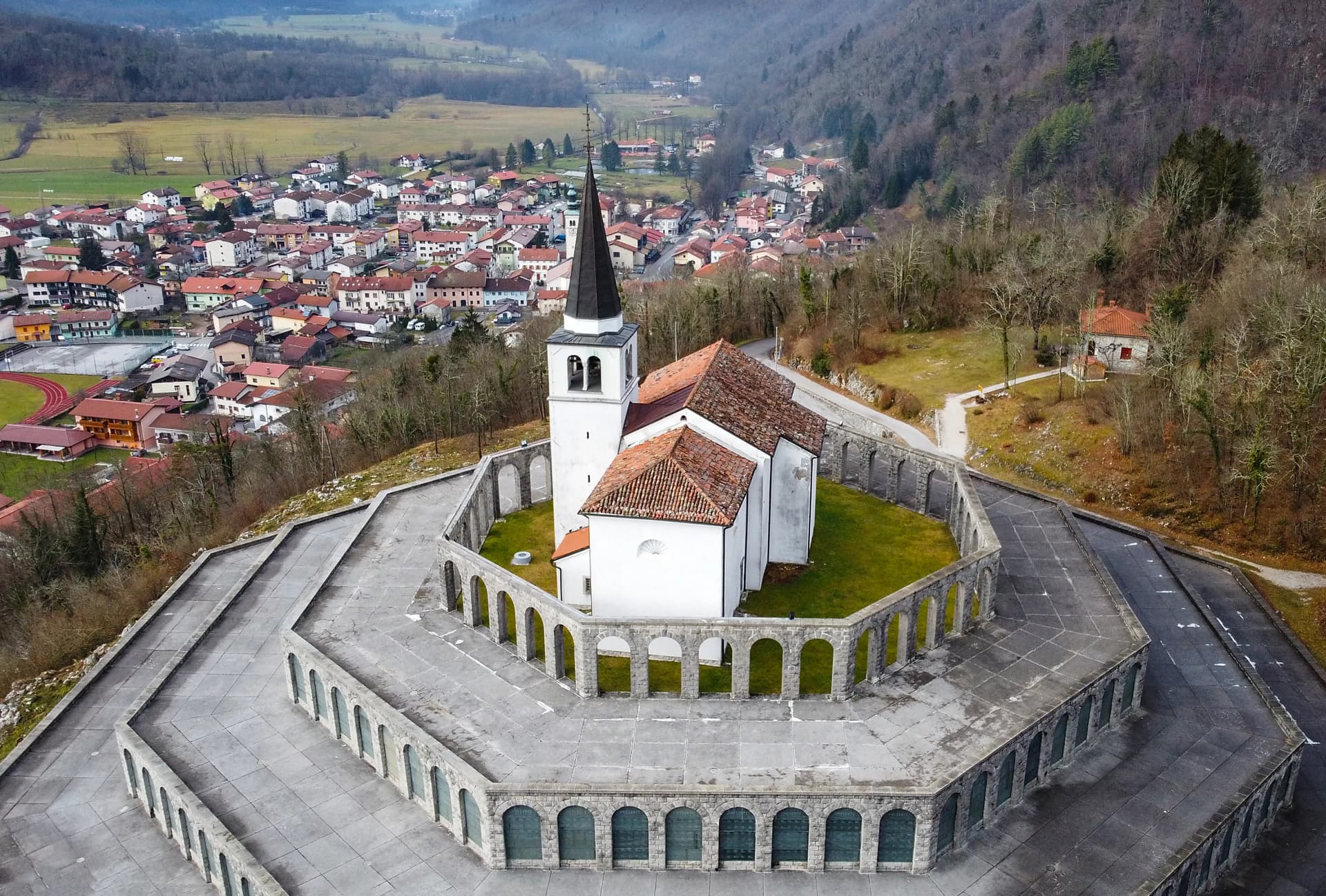 Aerial view of Italian military memorial with white church, stone arcade, and town below.