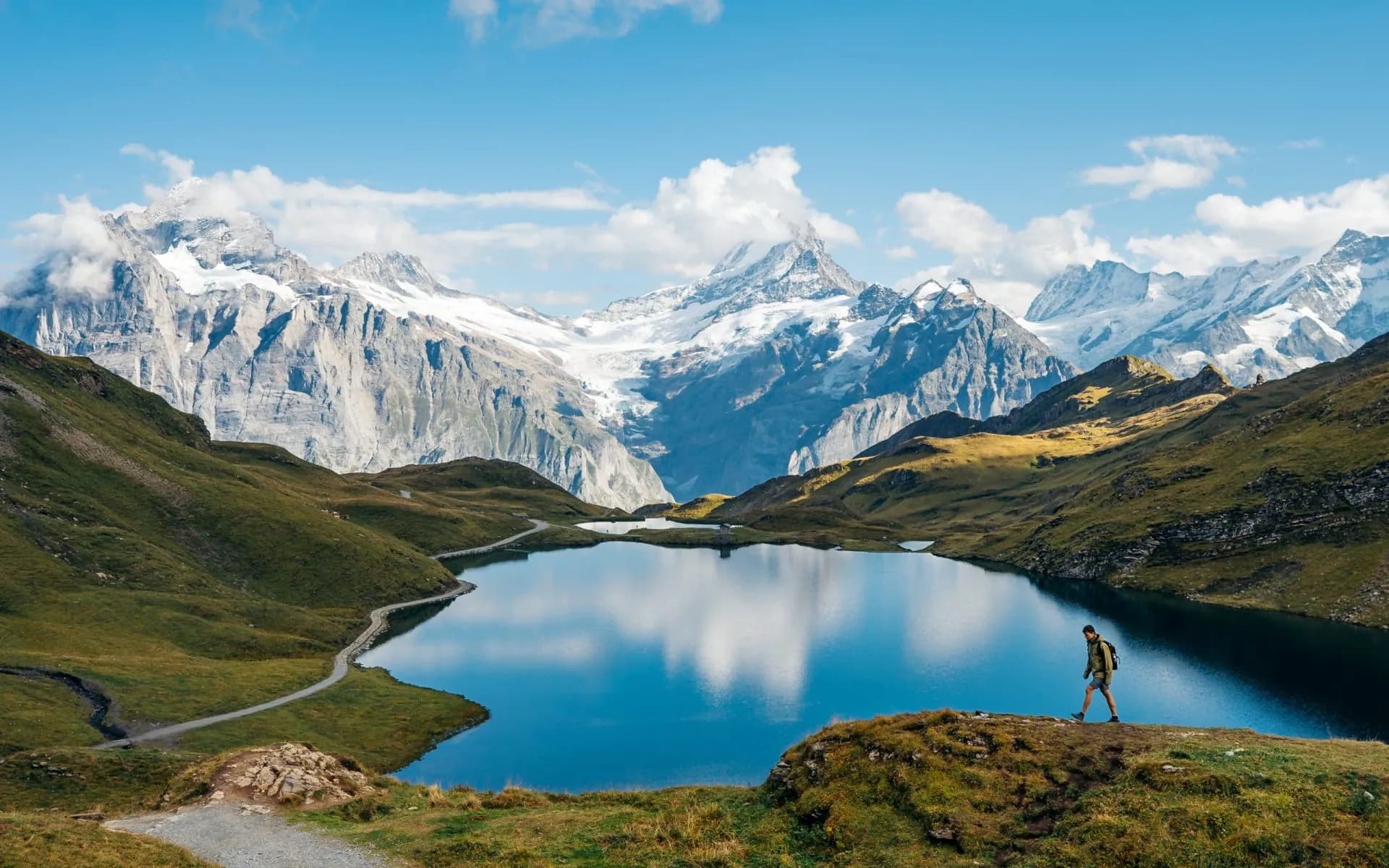 Hiker walking by Bachalpsee lake with reflection of snow-capped mountains under blue sky.
